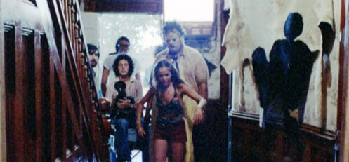 a behind the scenes picture from the texas chainsaw massacre. leatherface is looming behind a fleeing pam, while the cameraman and crew stand to the side obscured by the stairs.